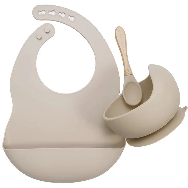 Beige silicone baby bib, bowl, and soft spoon tableware set on a white background